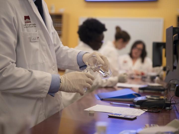 A student in lab coat pours compound ingredient out of a graduated cylindar into glass bottle