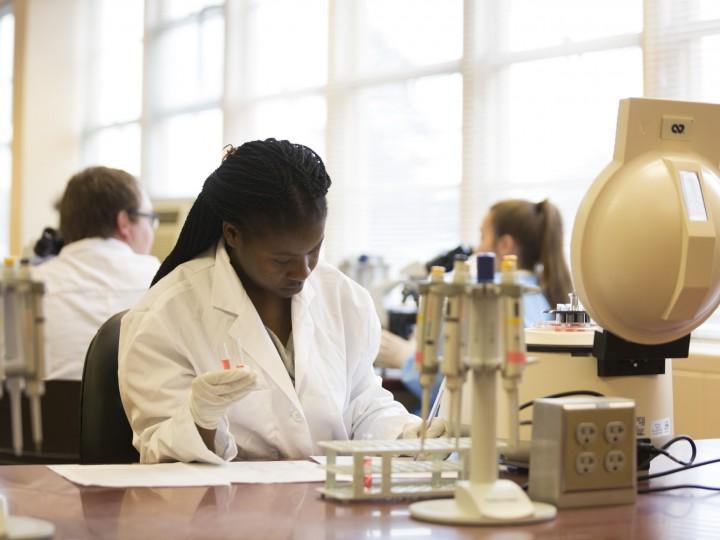 Student holding test tube with red liquid featuring pipette racks and benchtop centrifuge